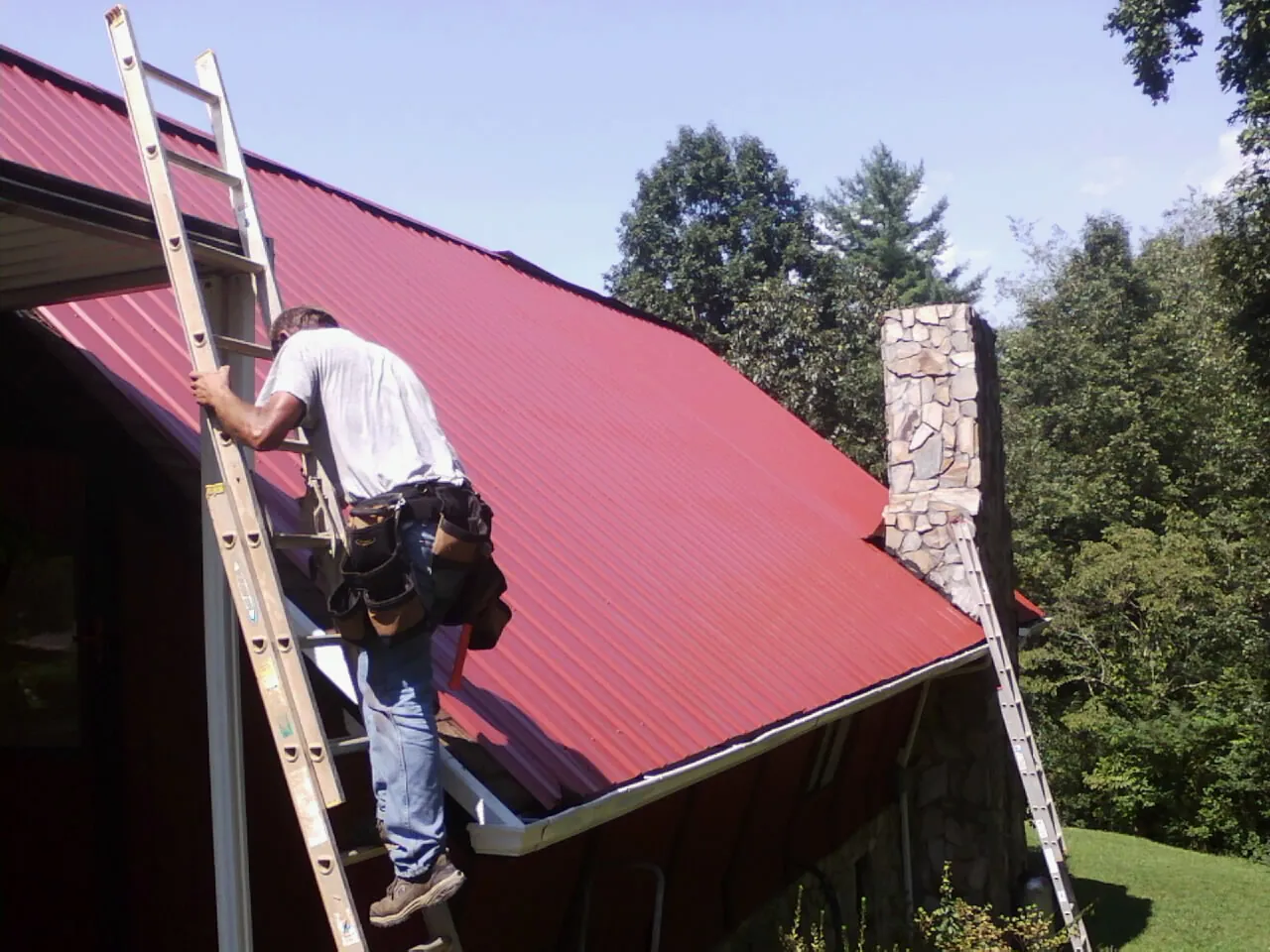 Roofer on a ladder beside a red metal roof and stone chimney — Soffit & Fascia Repair by Roofing Brothers in Munson